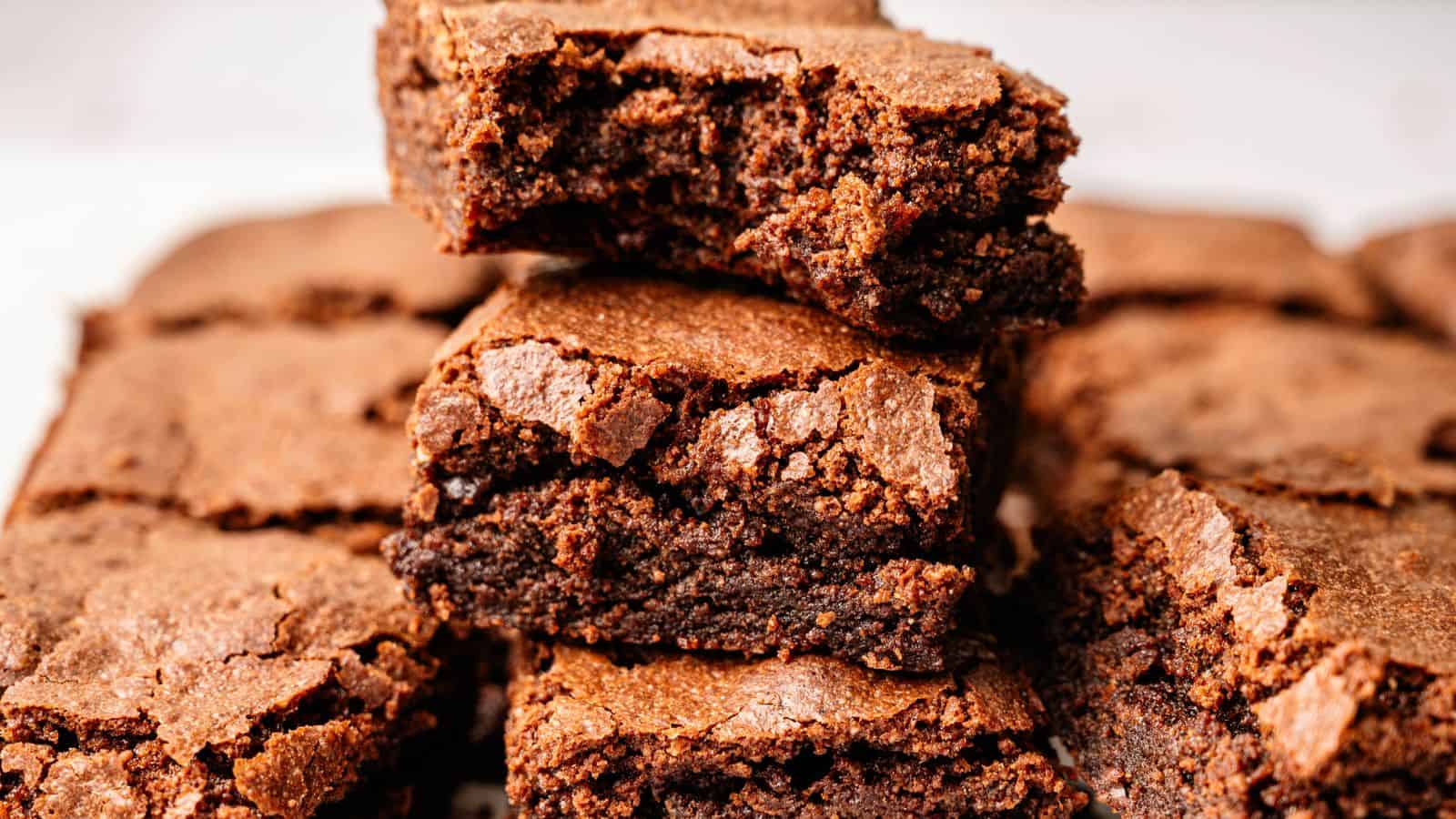 A stack of chocolate brownies with a cracked, crispy top rests on a surface, with more brownies arranged in the background.