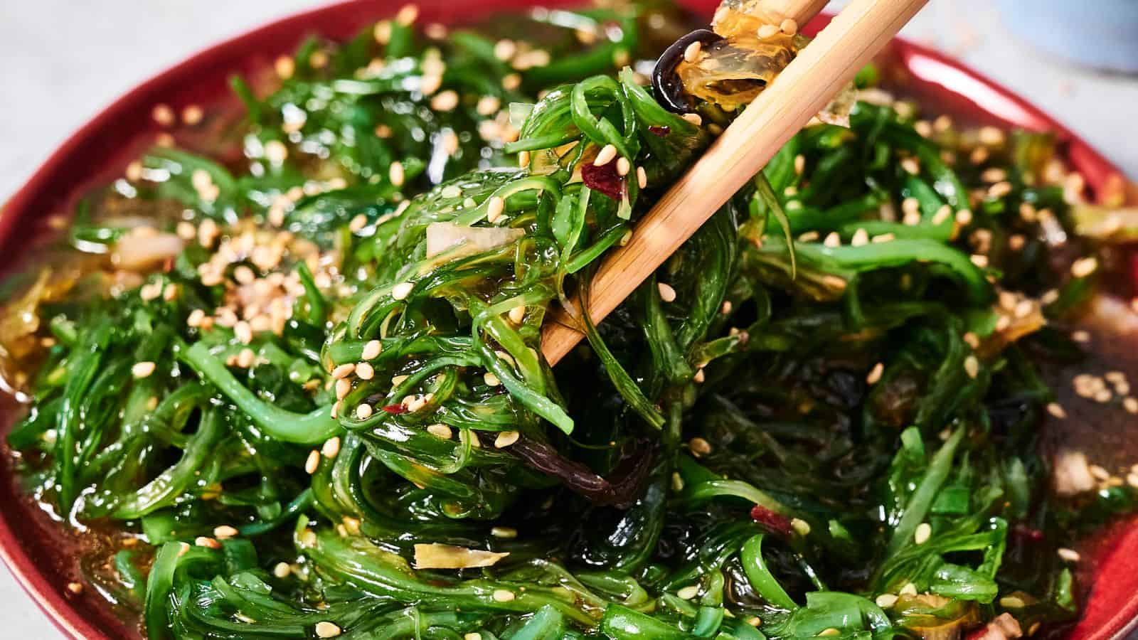 A close-up of seaweed salad with sesame seeds on a red plate, with chopsticks picking up a portion.