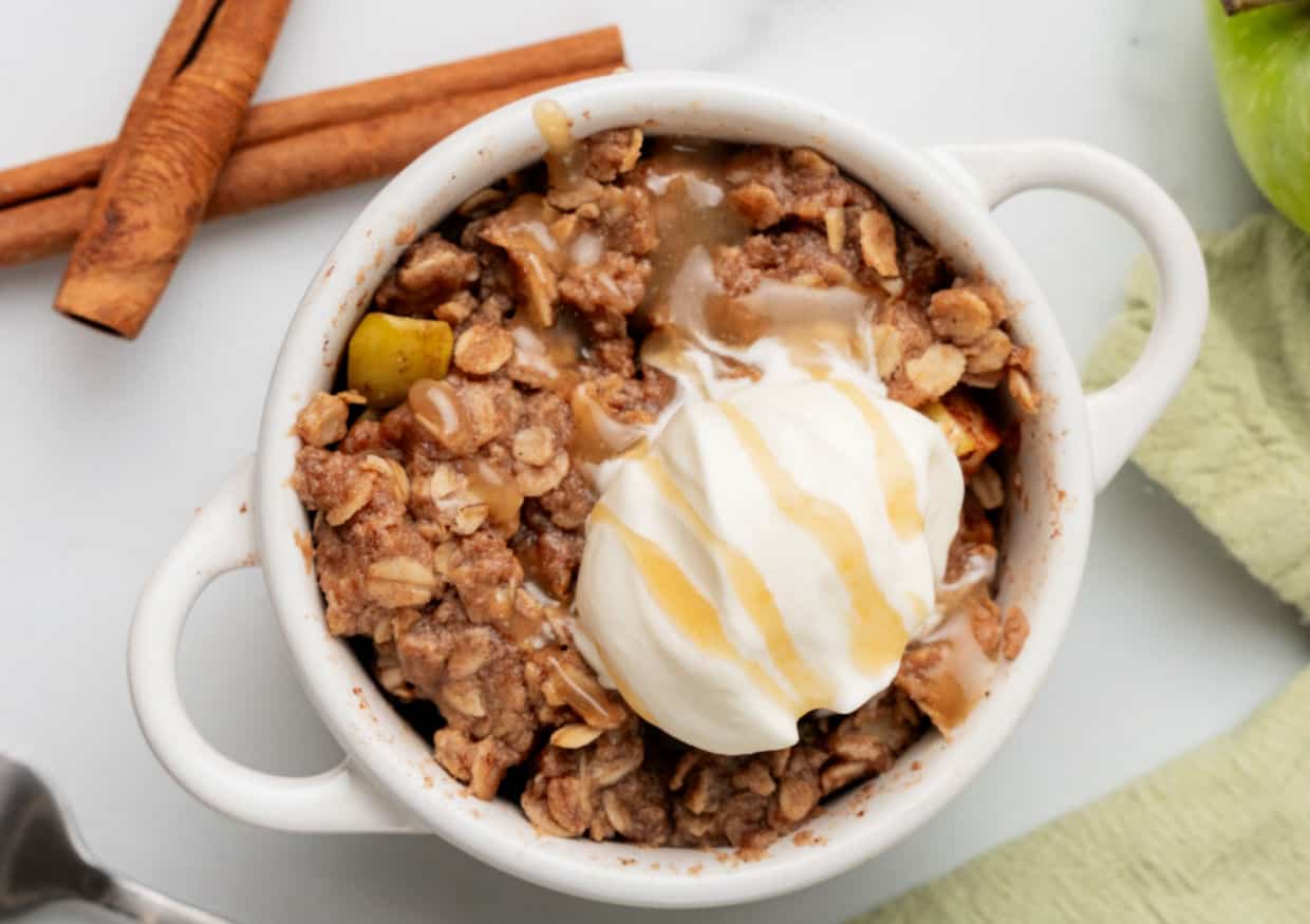 A white bowl of apple crisp topped with a scoop of vanilla ice cream and caramel drizzle, with cinnamon sticks and a green apple nearby.
