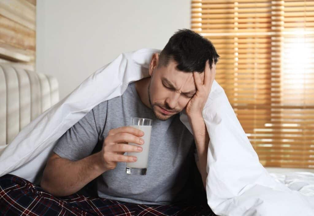 A man in pajamas sits on a bed under a blanket, holding his head with one hand and a glass of milk in the other, appearing tired or unwell—perfectly capturing the mood of national hangover day.