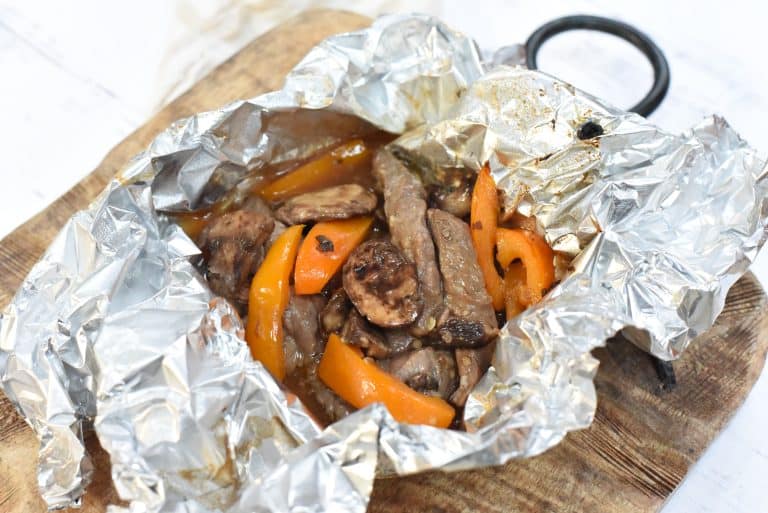 A serving of beef strips, mushrooms, and orange bell peppers cooked in foil, placed on a wooden board.