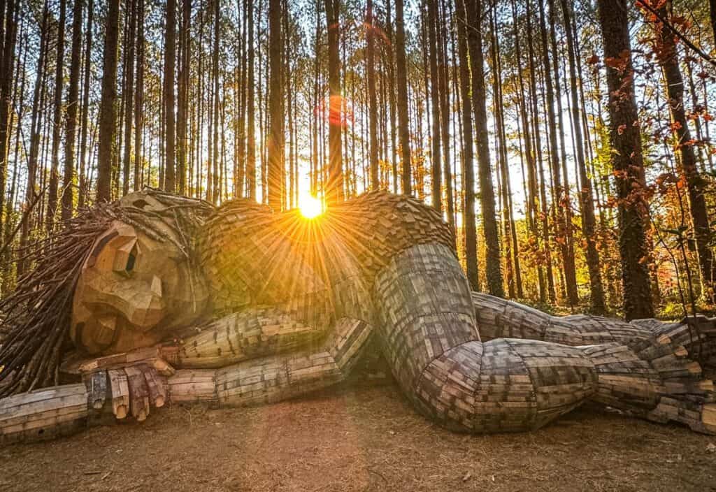 Large wooden sculpture of a reclining figure lies in a sunlit forest, with sunlight streaming through tall trees in the background.