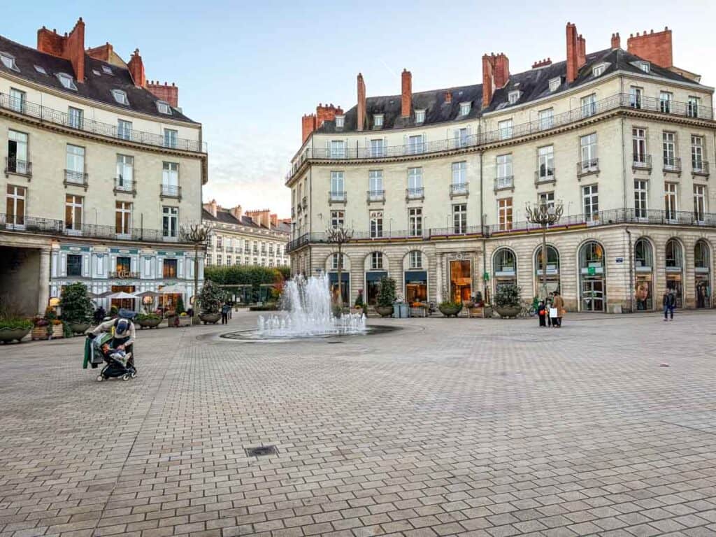 A paved city square with a central fountain, surrounded by multi-story buildings with shops on the ground floor; a few people are walking or sitting, capturing the vibrant atmosphere perfect for spending 48 hours in Nantes.