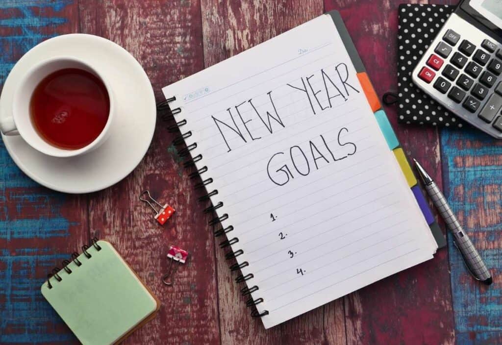 A notebook open to a page titled "New Year Goals" sits on a table with tea, a small notepad, pens, binder clips, and a calculator.