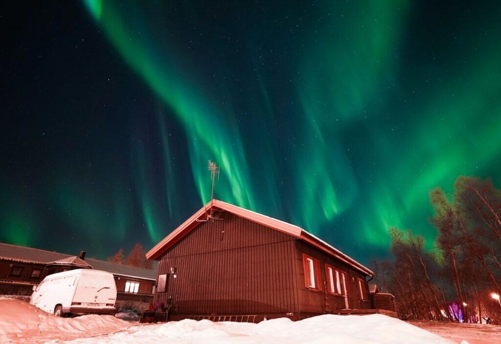 A snow-covered wooden house and van under a night sky illuminated by green northern lights.
