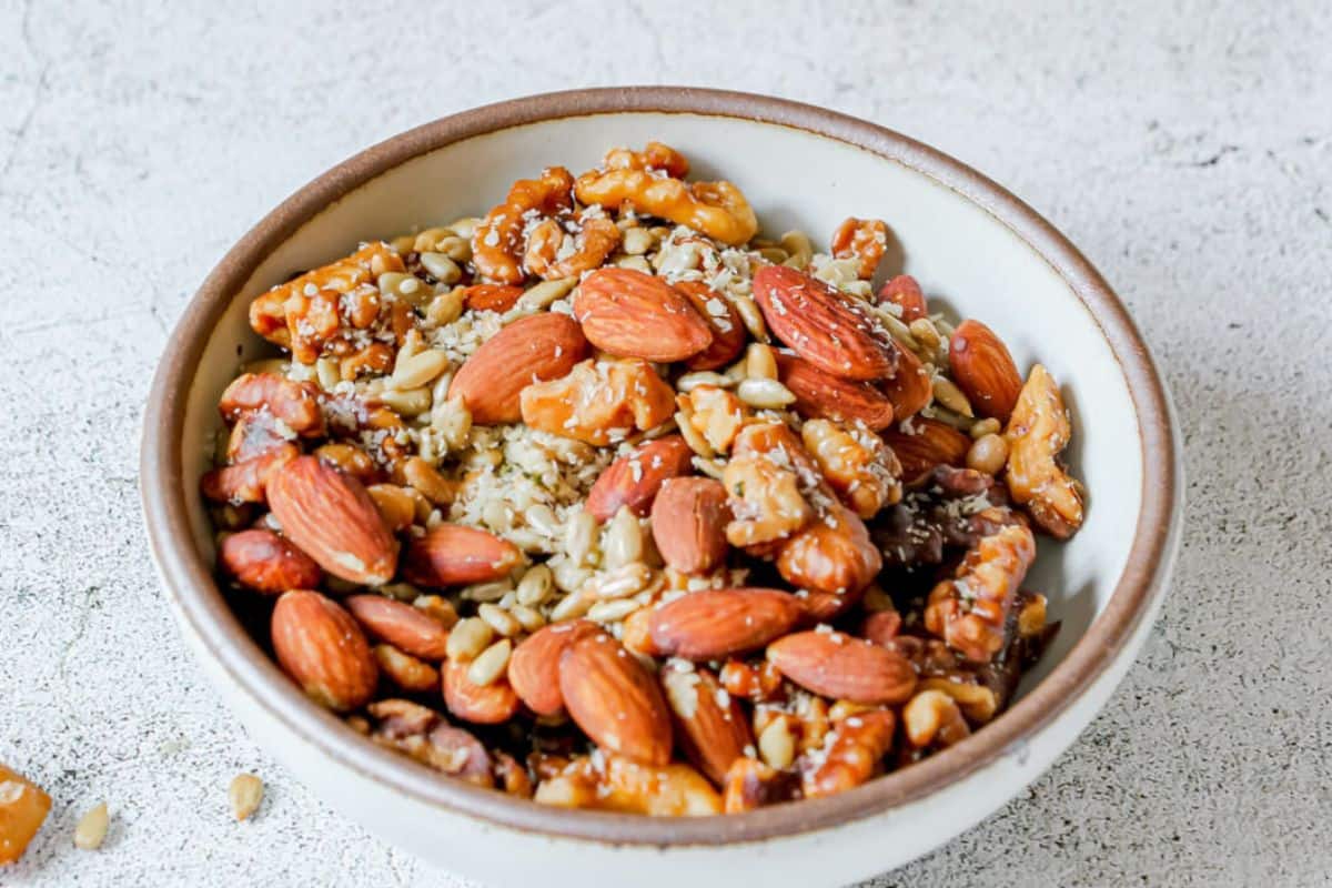 A bowl filled with a mix of almonds, walnuts, sunflower seeds, and other assorted nuts and seeds on a light textured surface.