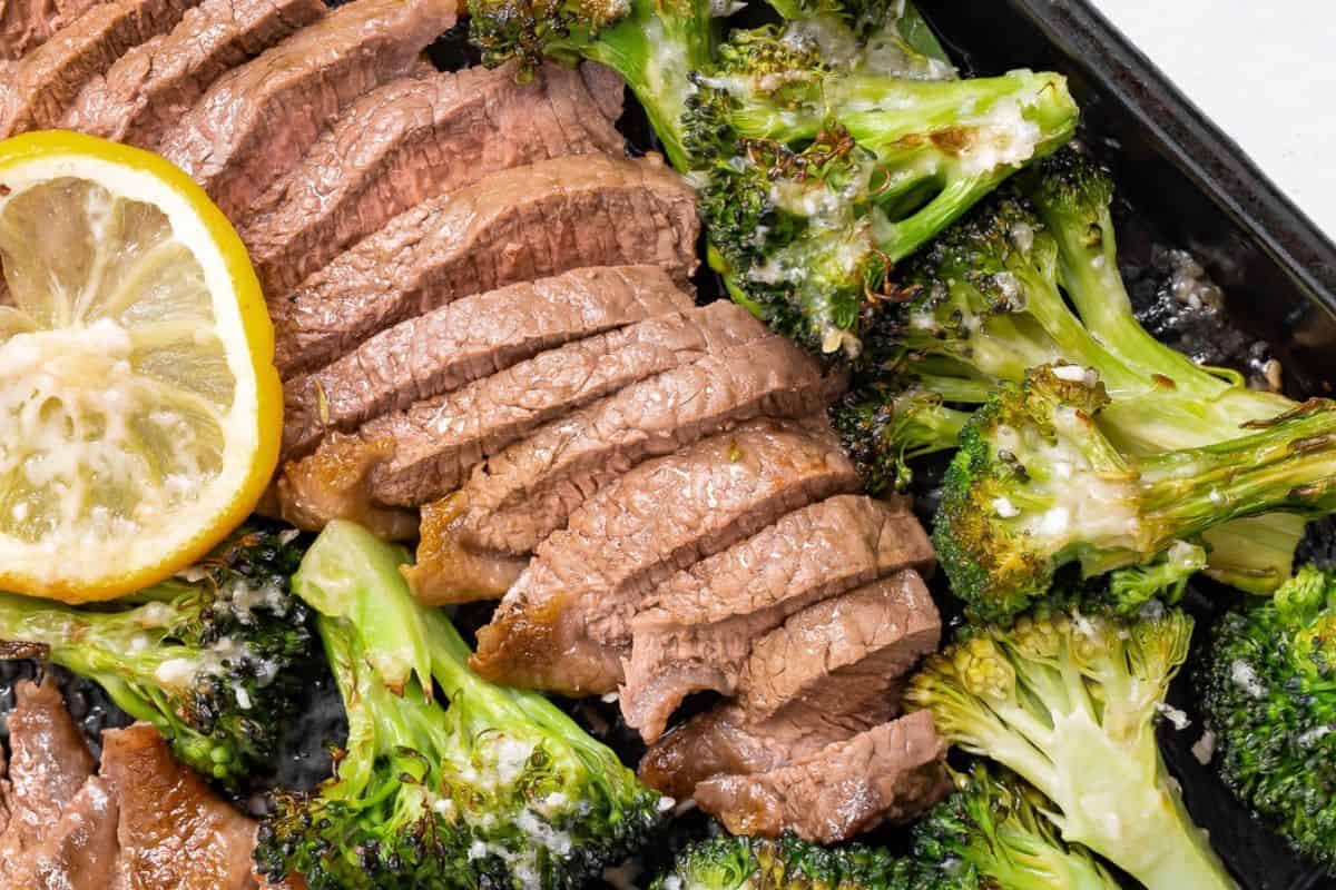 Sliced cooked beef arranged on a tray with roasted broccoli florets and a slice of lemon.