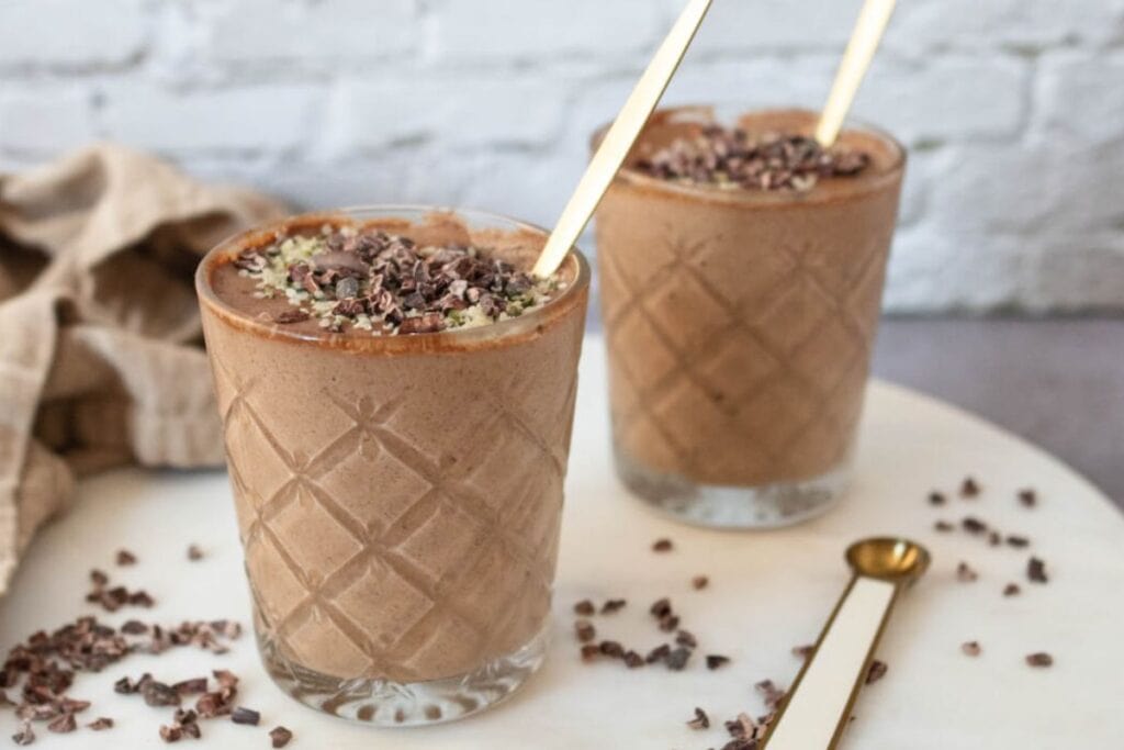 Two glasses filled with chocolate smoothie topped with cacao nibs and seeds, each with a gold spoon, placed on a white surface.