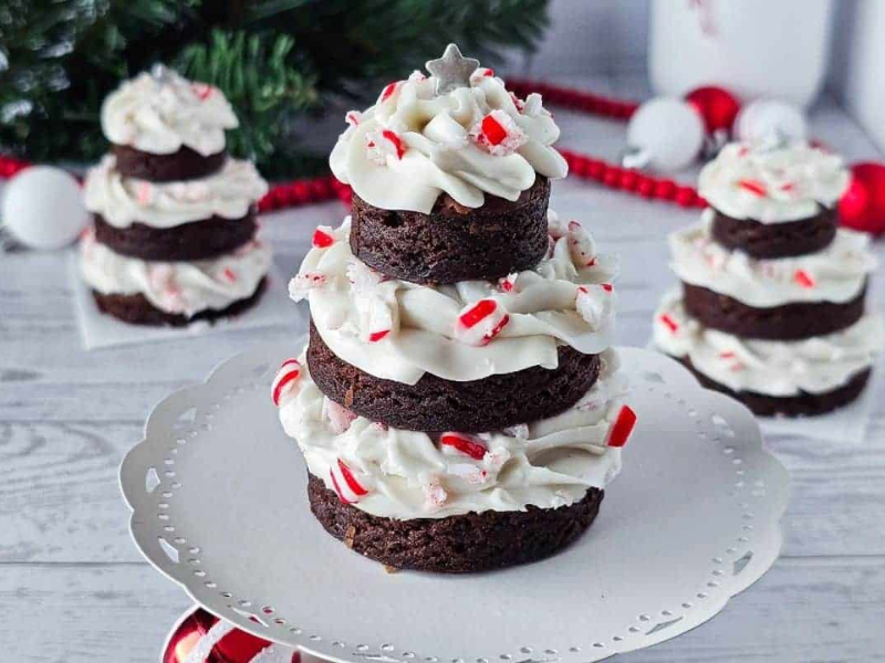 Three-tiered chocolate brownie dessert with white frosting and crushed peppermint, decorated to resemble a Christmas tree, displayed on a white cake stand with festive decor in the background.