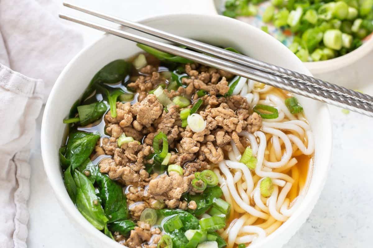 A bowl of noodle soup topped with cooked ground meat, chopped green onions, and leafy greens, with metal chopsticks resting on the rim.