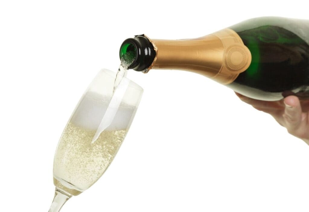 A hand pours sparkling wine from a green bottle with gold foil into a flute glass, creating lively bubbles and foam.