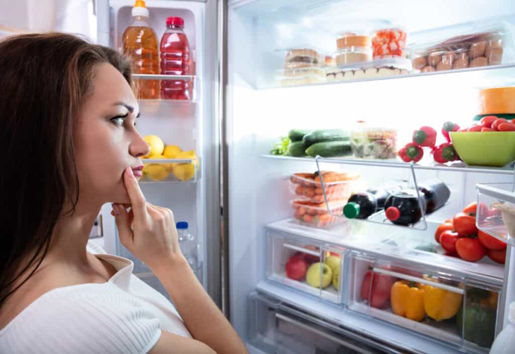 A woman stands in front of an open refrigerator, thoughtfully looking at shelves filled with various fruits, vegetables, and beverages.