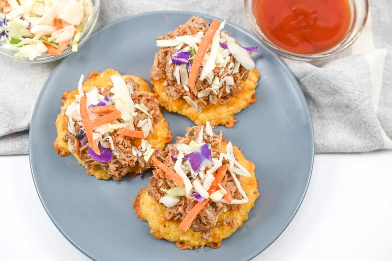 Three savory patties topped with shredded meat, coleslaw, and sliced carrots on a gray plate, with a bowl of dipping sauce and extra coleslaw on the side.