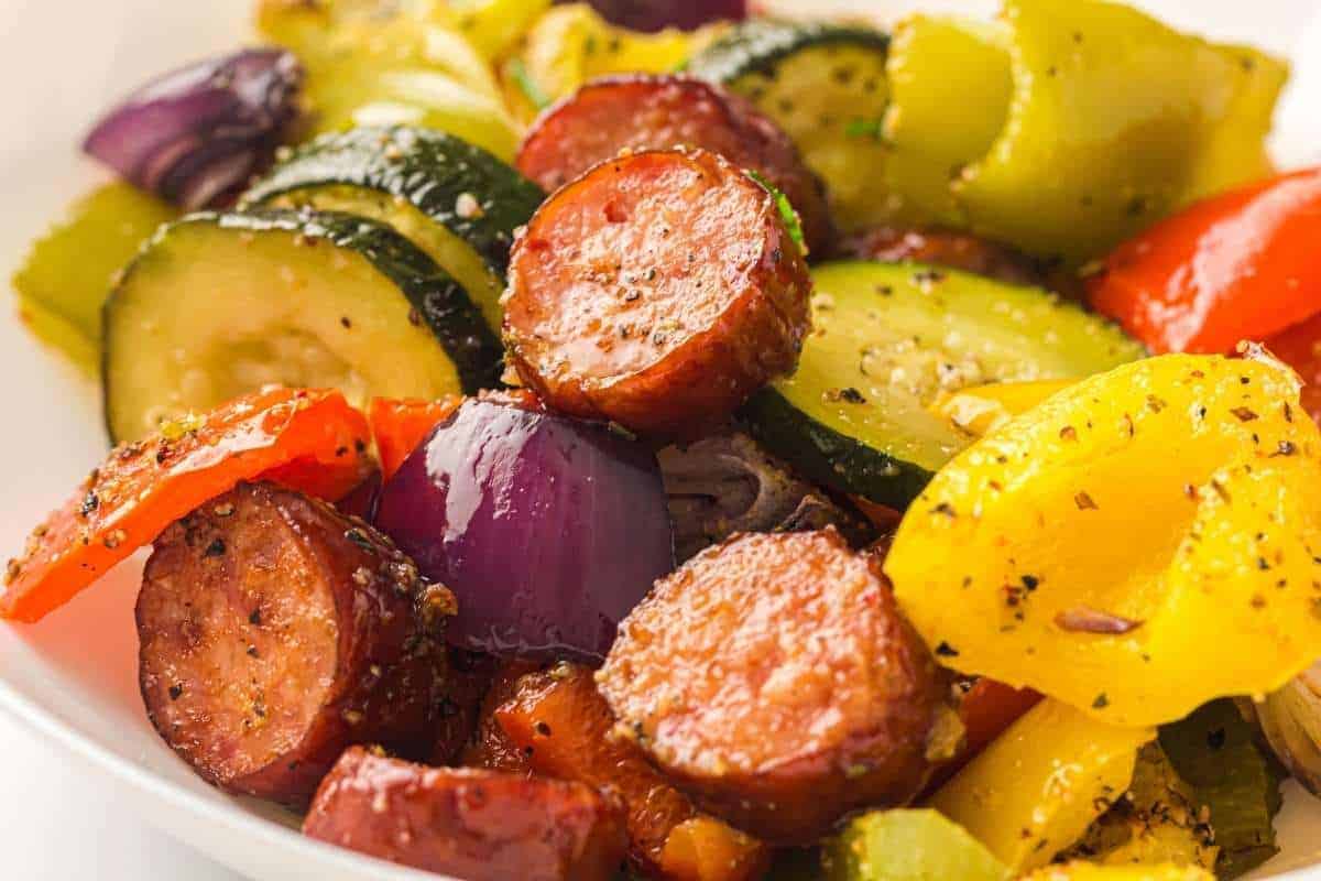 Sliced sausage, zucchini, red onion, yellow and red bell peppers seasoned with pepper and herbs on a white plate.