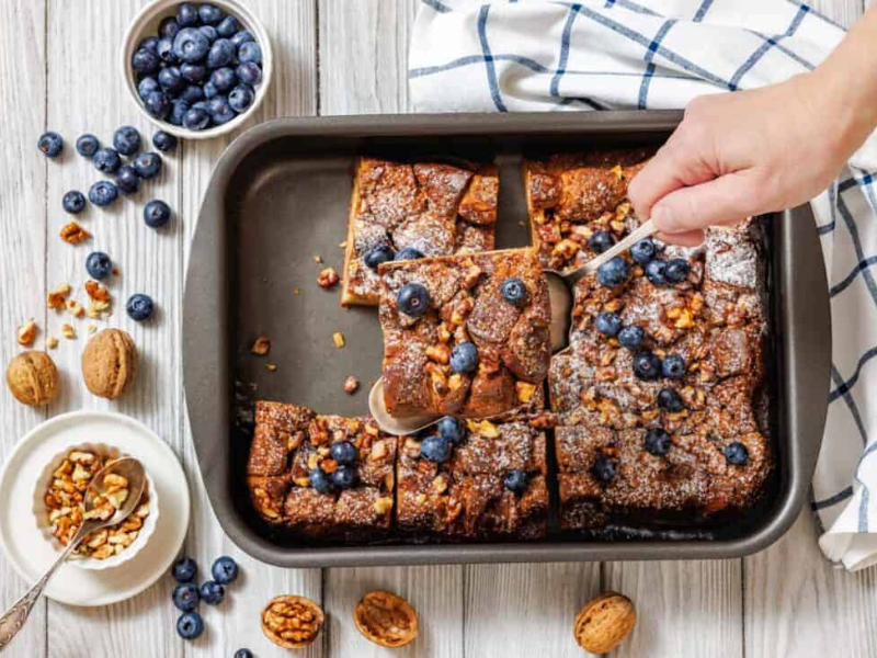 A baking pan with sliced Baked Cinnamon French Toast is shown, with a hand using a spoon to lift one piece.