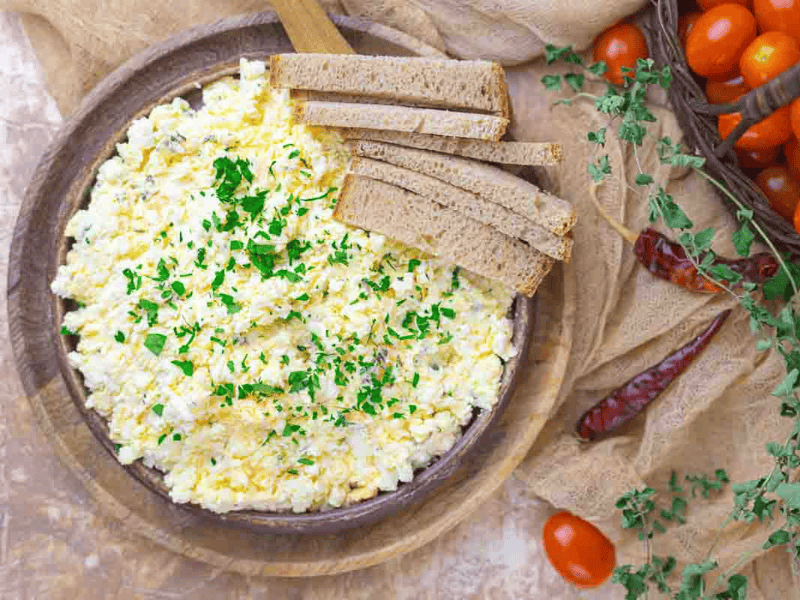 A plate of egg salad garnished with chopped herbs, served with slices of bread. Fresh tomatoes, dried peppers, and herbs are placed beside the plate.