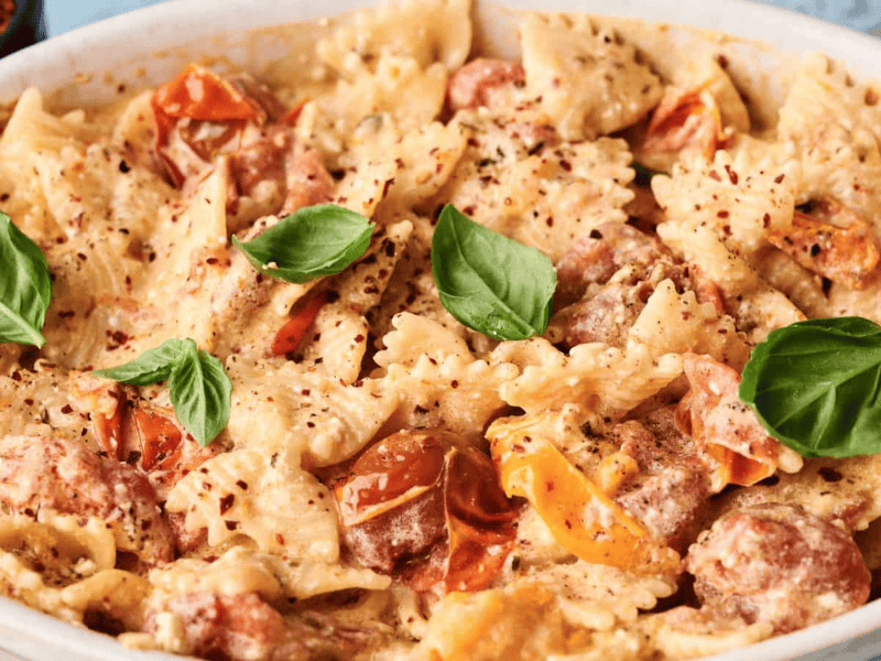 Baked bow-tie pasta with cherry tomatoes, creamy sauce, basil leaves, and red pepper flakes in a white dish.