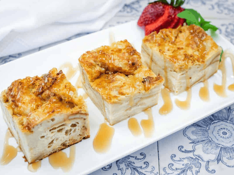 Three square pieces of Bourbon Croissant Bread Pudding with caramel drizzle are served on a white rectangular plate.