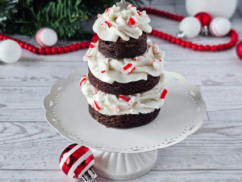 A three-tiered chocolate brownie cake with white frosting and red sprinkles sits on a white cake stand, surrounded by Christmas decorations.