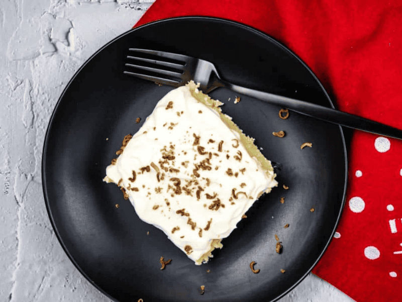 A square piece of cake with white frosting and chocolate shavings on a black plate, next to a black fork and a red cloth.