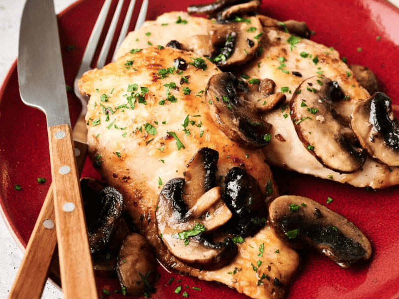 Two pieces of cooked chicken breast topped with saut&eacute;ed mushrooms and chopped herbs on a red plate, with a fork and knife beside them.
