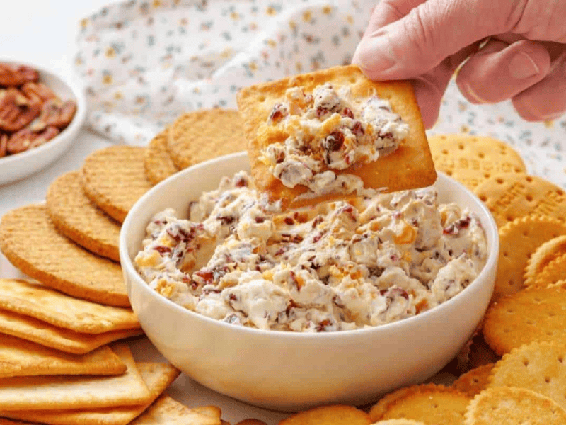 A hand dips a cracker into a bowl of cranberry cheese spread, surrounded by assorted crackers on a plate.