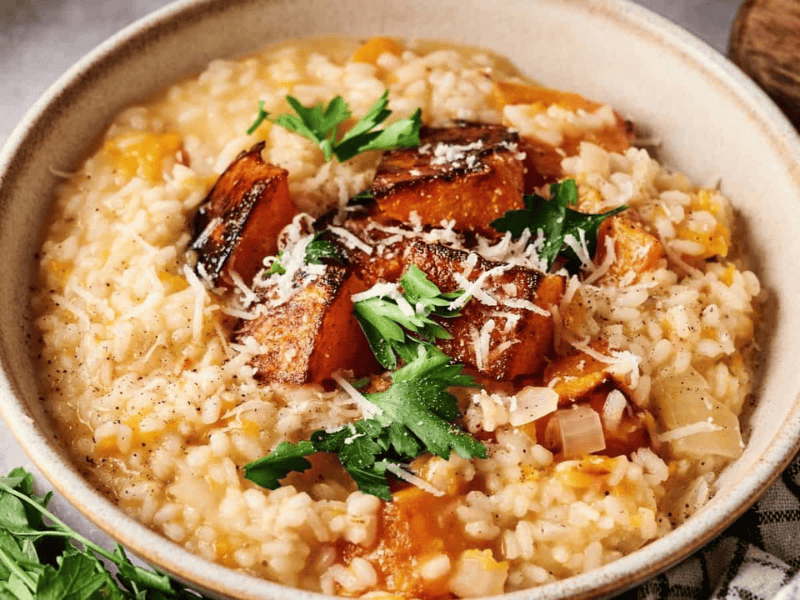 A bowl of risotto topped with roasted pumpkin pieces, grated cheese, and fresh parsley, with a checkered napkin and herbs nearby.