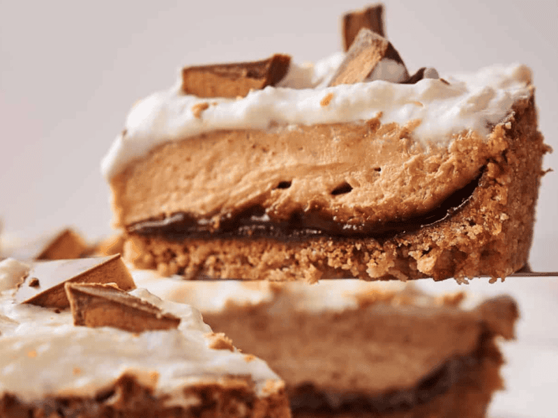 A slice of pie with a crumbly crust, chocolate layer, creamy peanut butter filling, whipped topping, and pieces of chocolate candy on top.