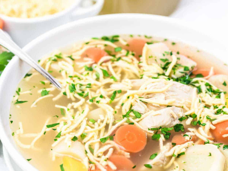A bowl of chicken noodle soup with shredded chicken, sliced carrots, noodles, and chopped parsley, with a spoon resting inside the bowl.