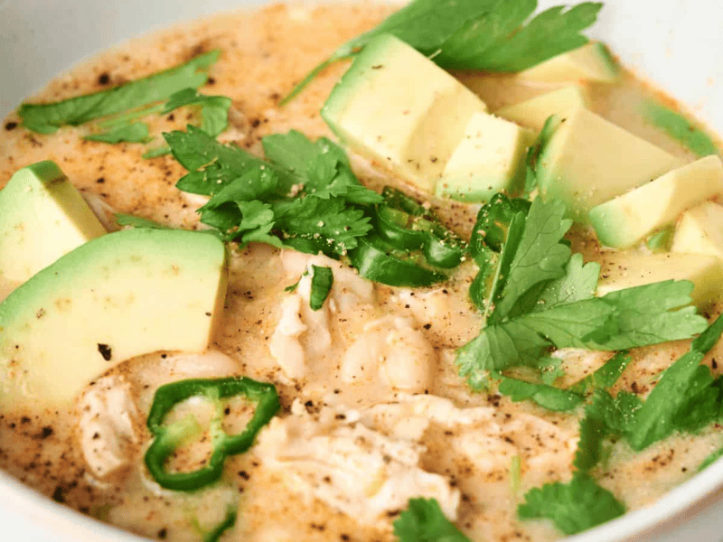 Close-up of a creamy soup with chunks of avocado, shredded chicken, white beans, sliced green chili, and fresh cilantro leaves.