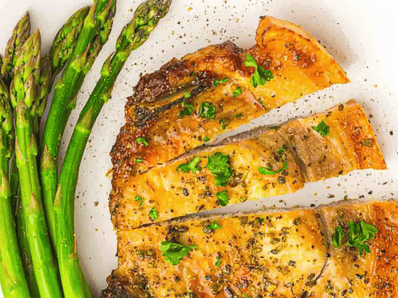 Cooked crispy pork chop garnished with herbs, served alongside steamed asparagus on a white plate.