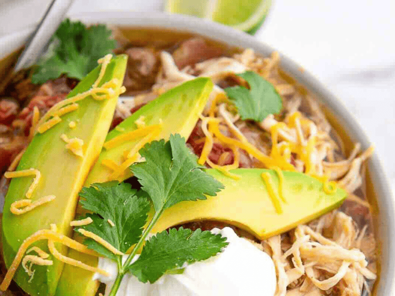 A bowl of shredded taco soup topped with sliced avocado, shredded cheese, cilantro, and a dollop of sour cream.