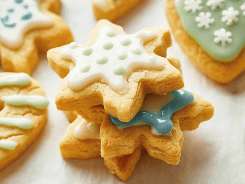A stack of star-shaped sugar cookies decorated with white and blue icing sits on a white surface, with more cookies in the background.