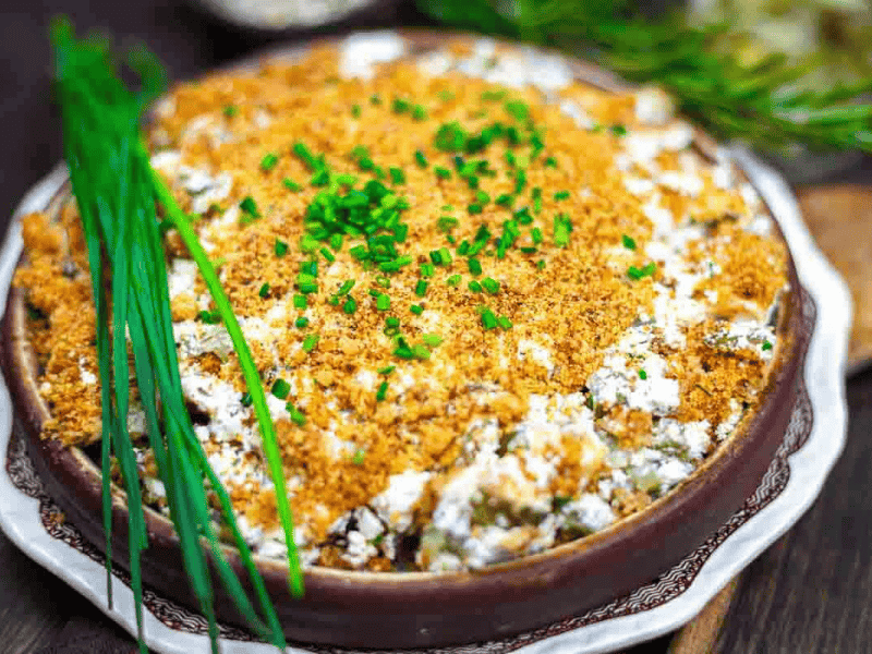 A baked dill pickle dip topped with golden breadcrumbs and chopped chives, garnished with fresh herbs on the side, served in a round dish.