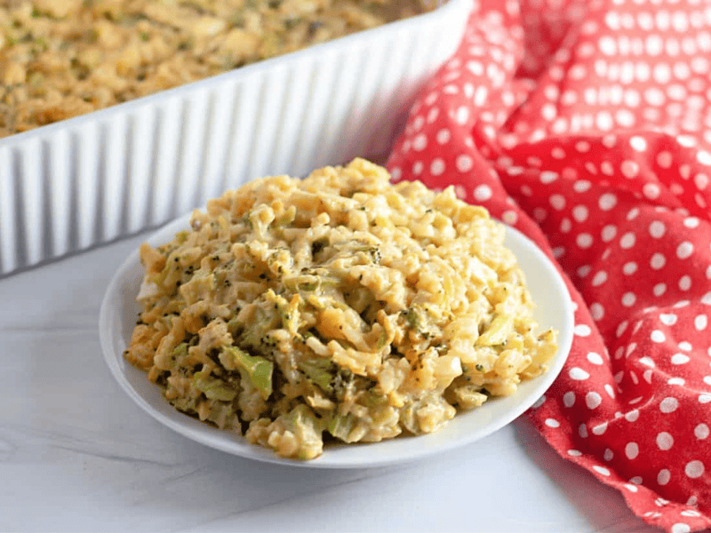 A plate of Easy Cheesy Broccoli Rice Casserole sits on a white surface next to a red and white polka dot cloth, with a baking dish in the background.