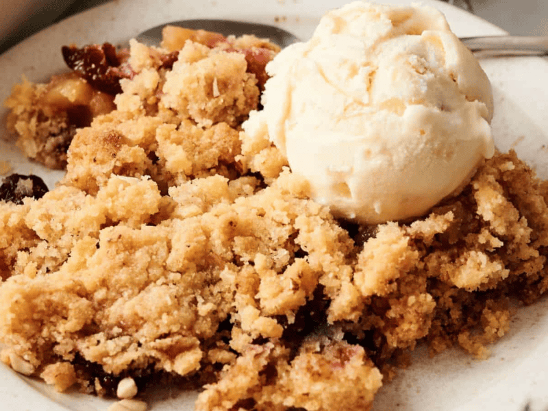 A plate of fruit crumble topped with a scoop of vanilla ice cream, accompanied by a spoon.