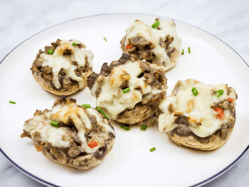 Five stuffed mushrooms topped with melted cheese are arranged on a white plate, garnished with chopped chives.