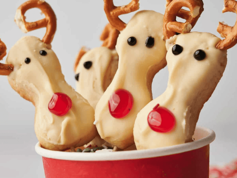 Three reindeer-shaped cookies with pretzel antlers, candy noses, and chocolate eyes are arranged in a red cup.