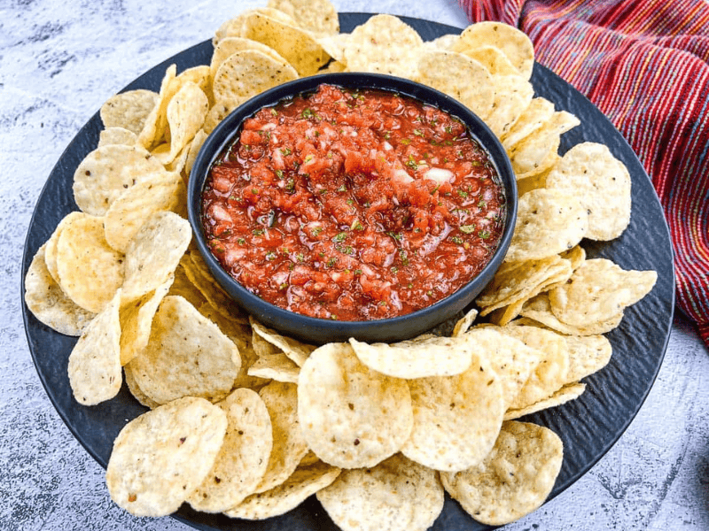 A black plate with round tortilla chips arranged around a bowl of chunky red salsa, placed on a light-colored surface with a red-striped cloth nearby.