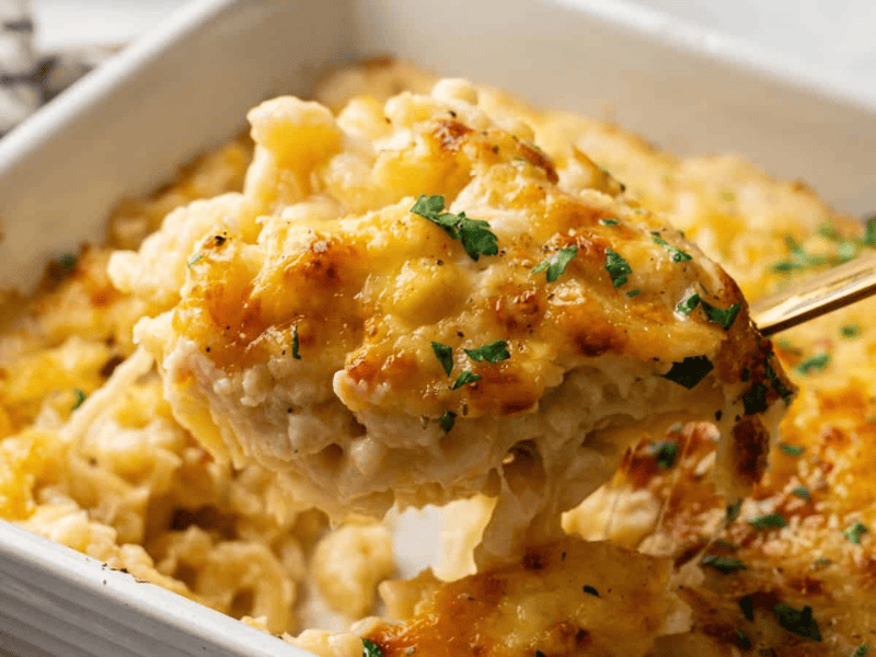 A close-up of a serving of baked macaroni and cheese being lifted from a casserole dish, topped with melted cheese and garnished with chopped herbs.