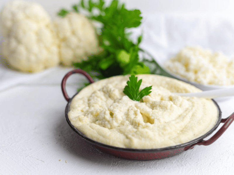 A bowl of creamy mashed cauliflower garnished with a parsley leaf, with raw cauliflower and parsley in the background.
