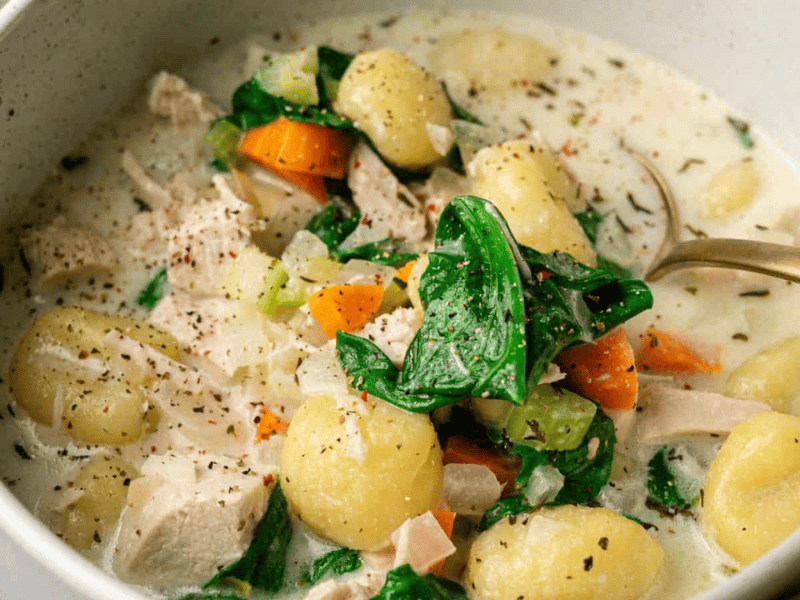 Creamy soup with gnocchi, spinach, carrots, celery, diced chicken, and herbs in a white bowl.