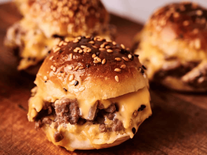 Three cheeseburger sliders with ground beef, melted cheese, and sesame seed buns are arranged on a wooden surface.