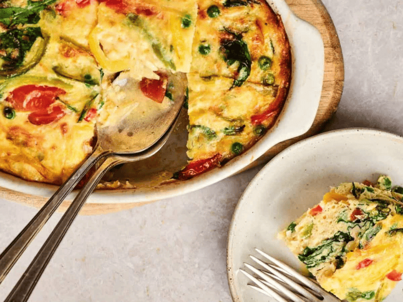 A vegetable frittata in a baking dish with a serving spoon, next to a plate holding a sliced portion of the frittata.