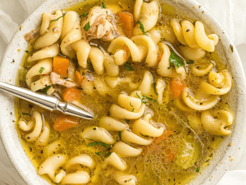 A bowl of rotini pasta soup with pieces of chicken, carrots, herbs, and broth, with a spoon resting inside.