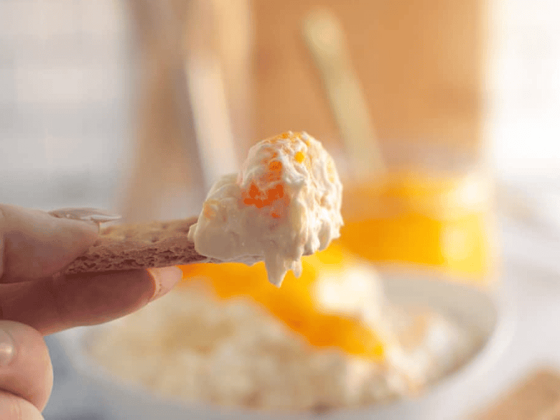 A hand holds a cracker topped with a creamy cheese spread and small pieces of orange fruit, with a bowl and jar in the blurred background.
