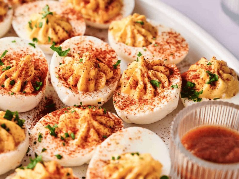 A plate of deviled eggs garnished with paprika and chopped parsley, with a small dish of sauce on the side.