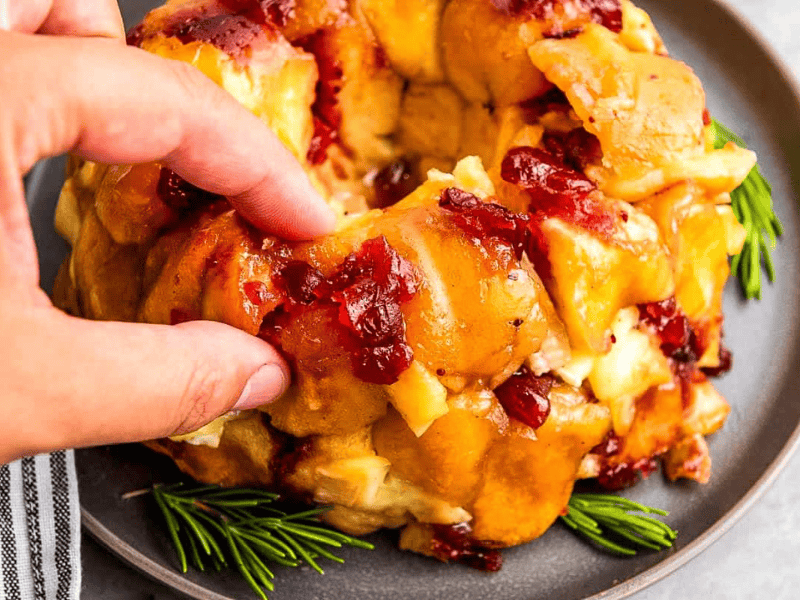 A hand pulls a piece from a baked pull-apart bread ring with visible cranberries, cheese, and herbs, served on a gray plate with rosemary garnish.