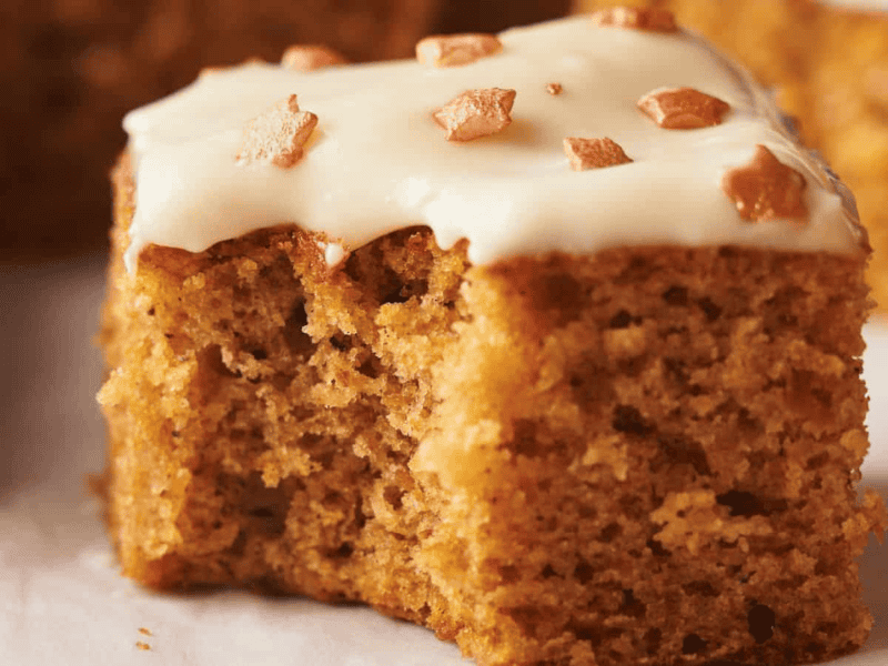 A close-up of a square slice of spiced cake topped with white icing and small brown sprinkles, showing a moist and airy texture.