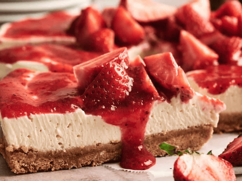 A slice of cheesecake with a graham cracker crust, topped with strawberry sauce and fresh strawberry pieces, sits on a serving tray.